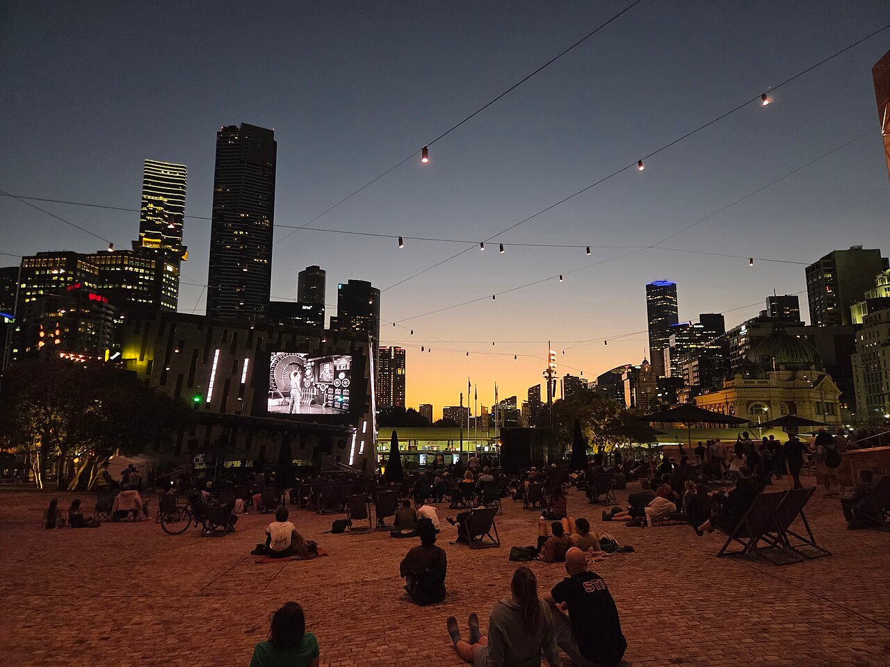 People sitting in Federation Square at dusk, with city buildings and strings of lights beyond.